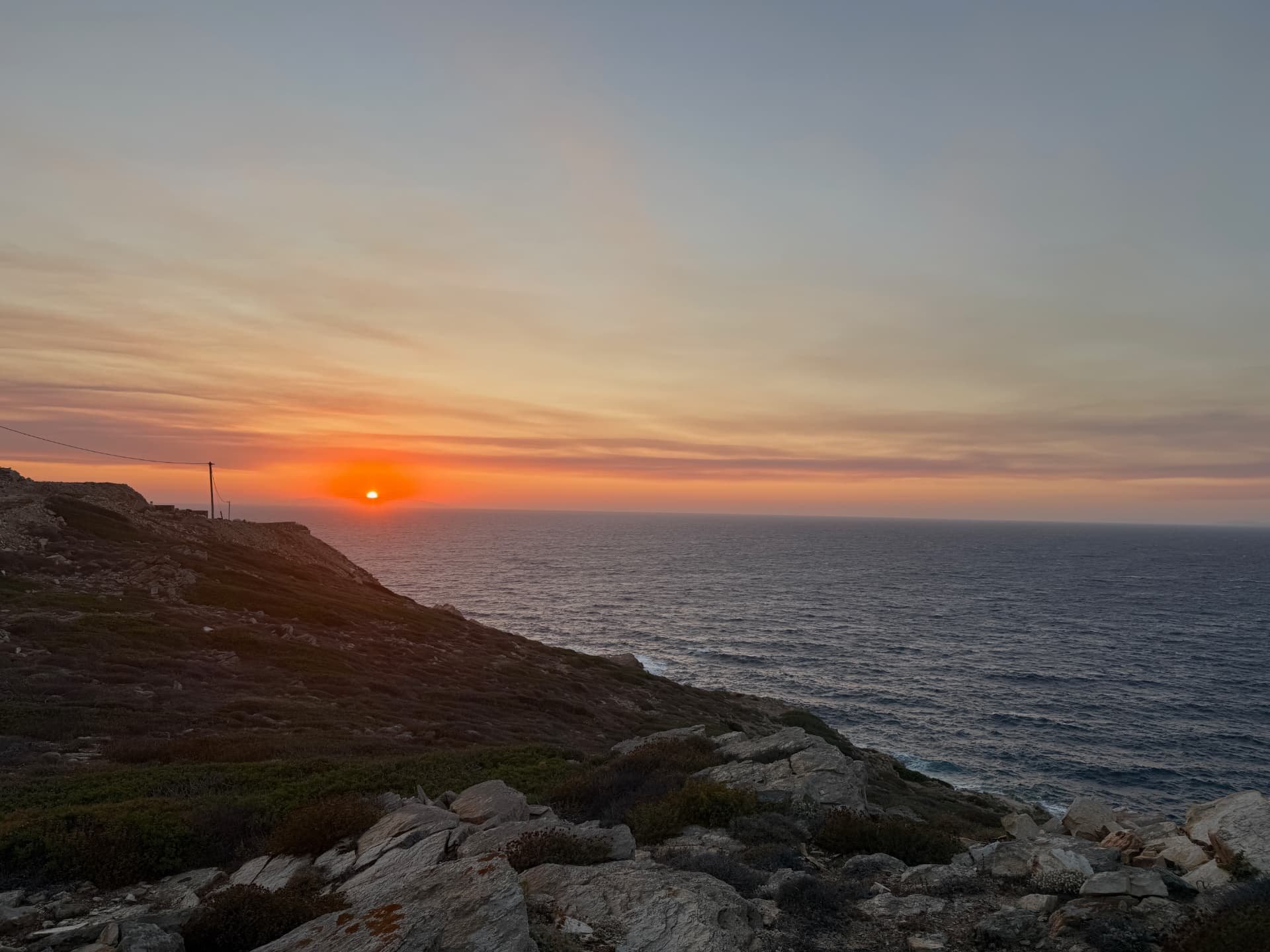 The sun setting over the open Aegean Sea beyond the rocky, low-vegetation landscape of the Akri House site.